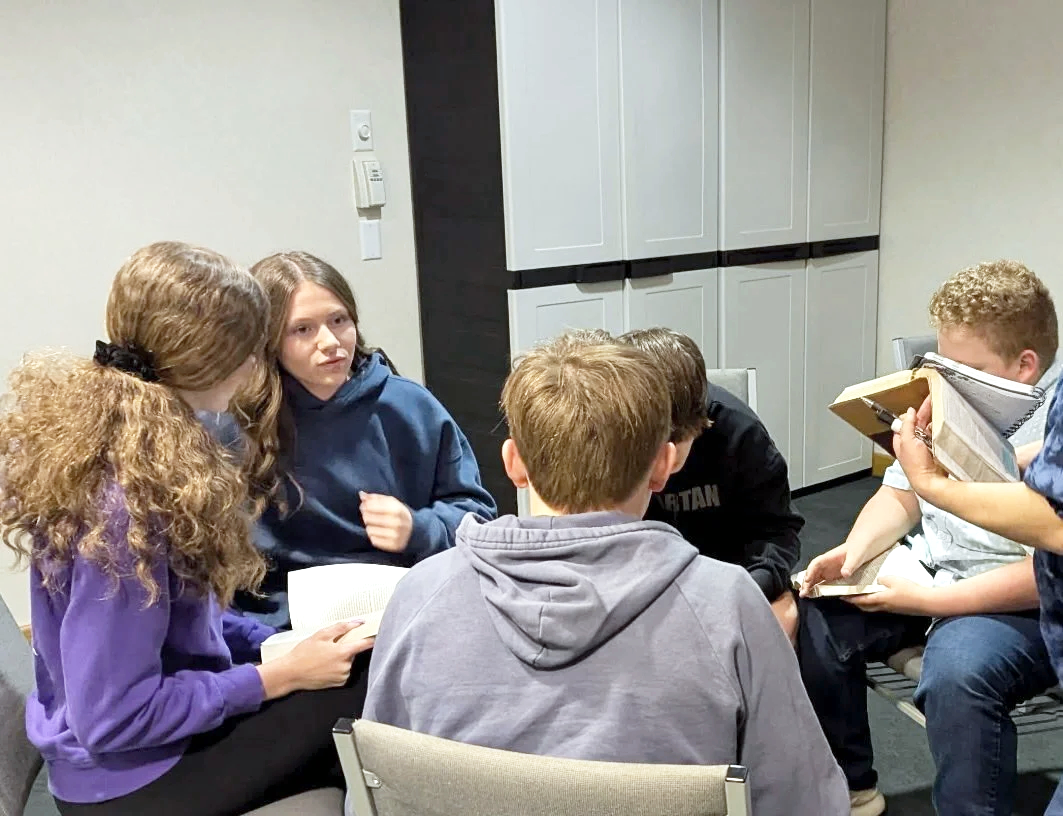 Group of six teenagers sitting in a circle having a discussion, some holding books, in a room with cabinets in the background.