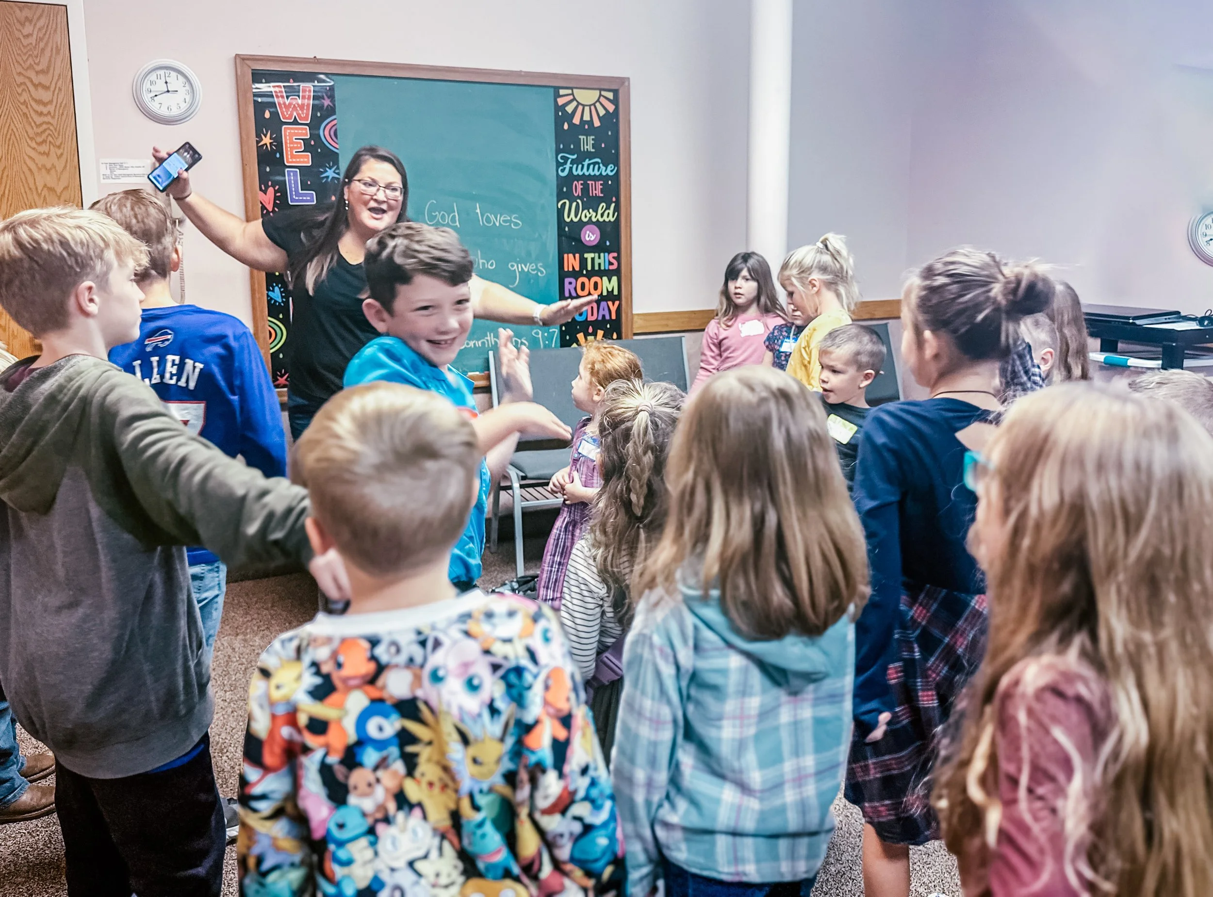 A classroom with a group of children gathered around a woman teaching at the front. The woman is smiling and holding a smartphone, and there are chalkboards with writing and colorful decorations behind her.