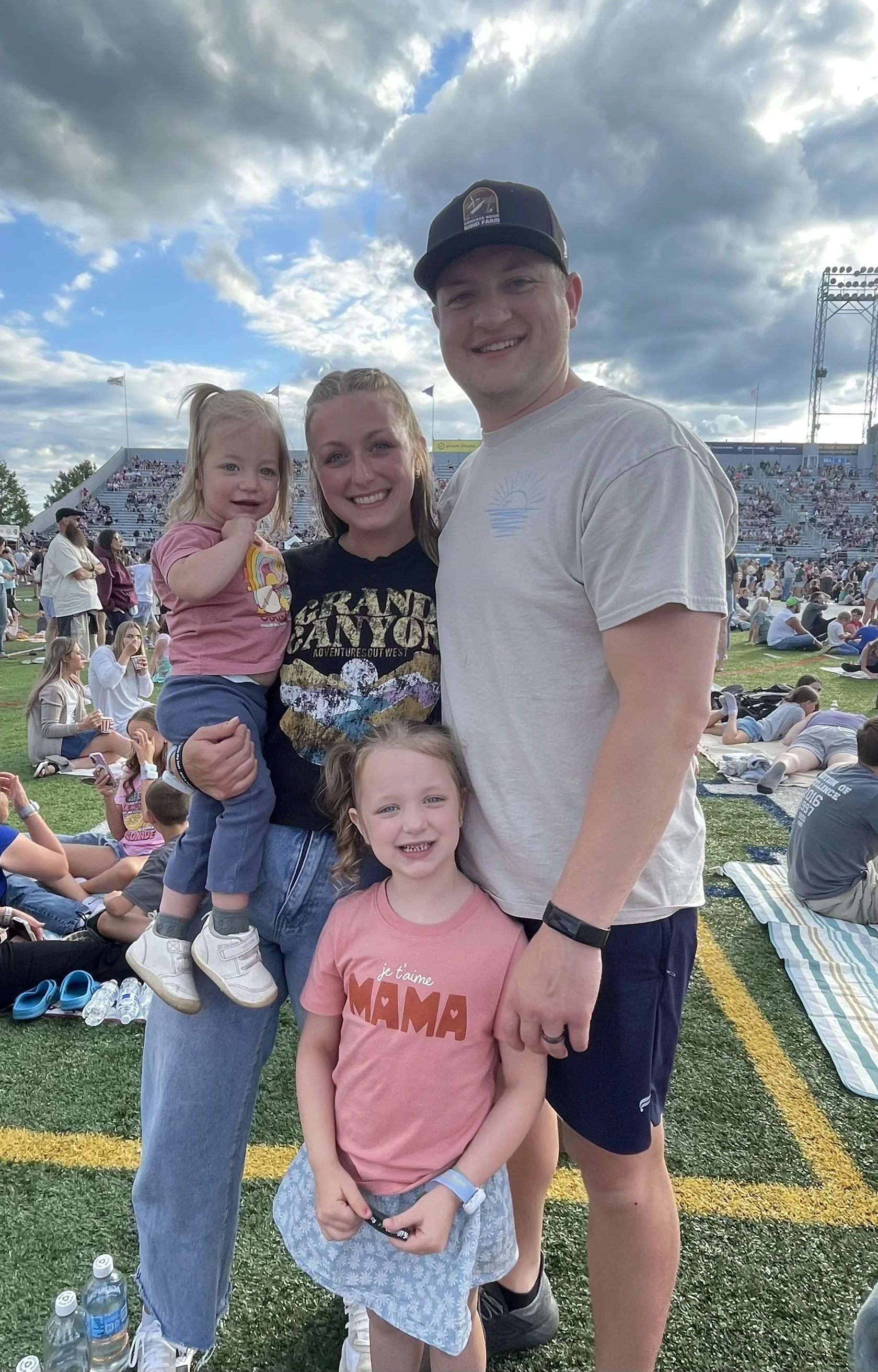 A group of four people, two children and two adults, smiling and posing together at an outdoor event with a large crowd, stadium seating, and a stage in the background. The sky is partly cloudy.