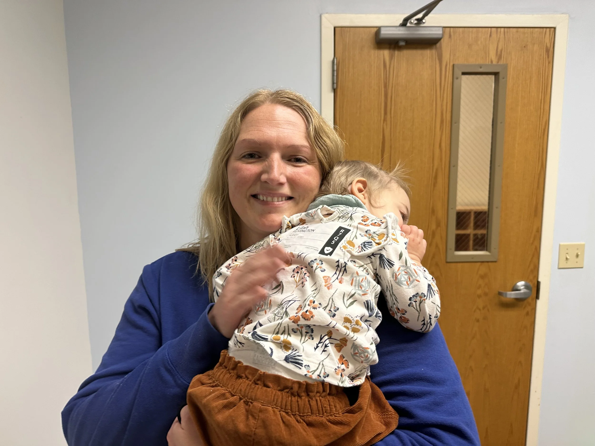 A woman with blonde hair smiling while holding a young child with blonde hair, wearing a white floral shirt and brown pants, against a door and white wall background.