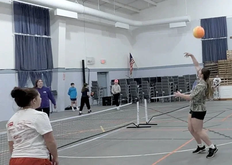 A girl is playing volleyball in an indoor gymnasium, with several other children in the background, along with chairs stacked against the wall and an American flag. The girl is preparing to bump the volleyball over the net.