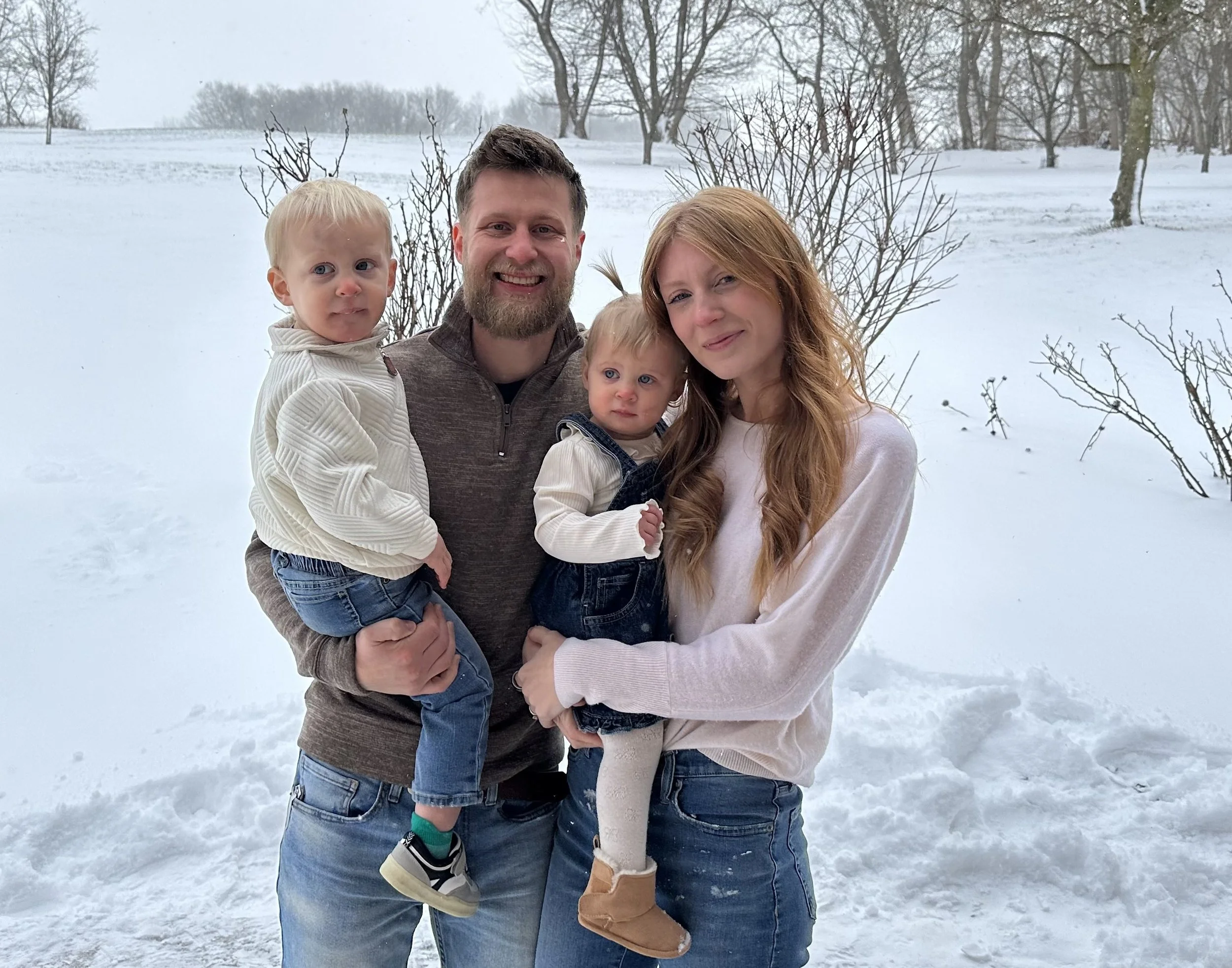 A family of four standing outdoors in snow-covered landscape with trees.