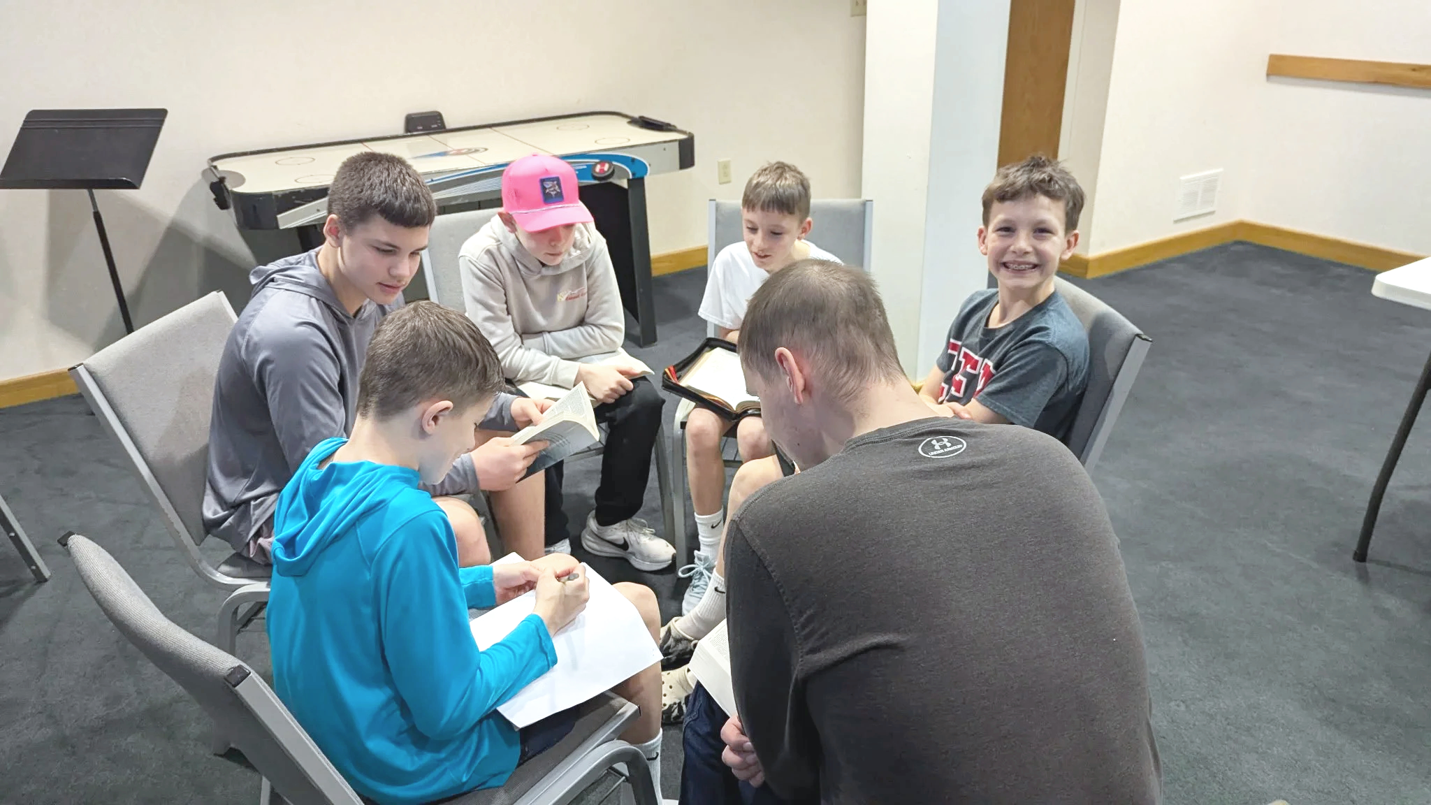 A group of seven kids and one adult sitting in a circle in a room, reading or writing, with a shuffleboard table in the background.