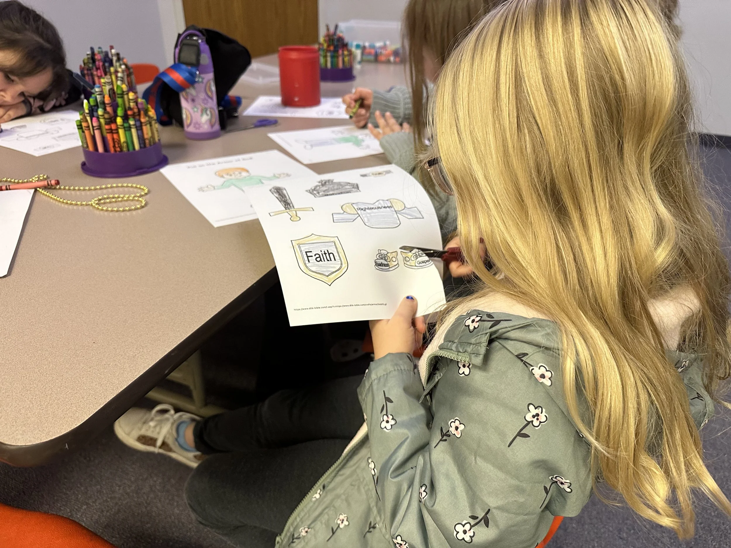 A little girl with long blonde hair wearing glasses and a green jacket with white flowers, sitting at a table and drawing on a coloring sheet with images of a shield labeled "Faith" and other symbols. Other children are at the table with art supplies and drawings.