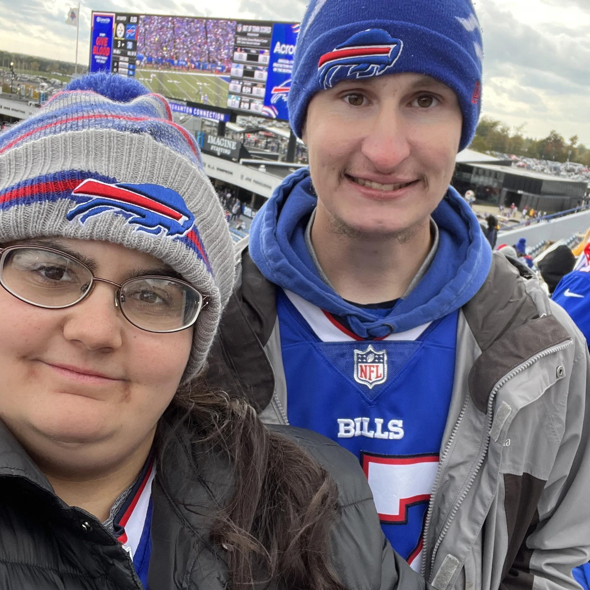 Two fans wearing Bills hats and jerseys at a football game, with a stadium and a large screen in the background.