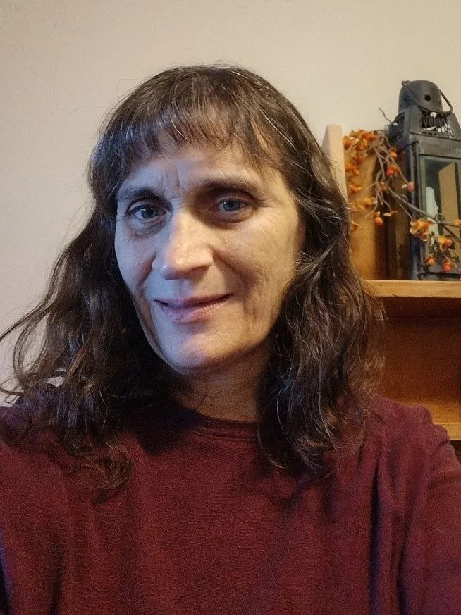 A woman with shoulder-length brown hair and a maroon shirt taking a selfie indoors, with a wooden shelf and some decorative items in the background.