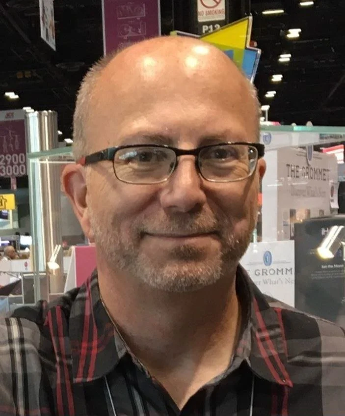 A middle-aged man with glasses, short hair, and a beard taking a selfie at a trade show or convention.