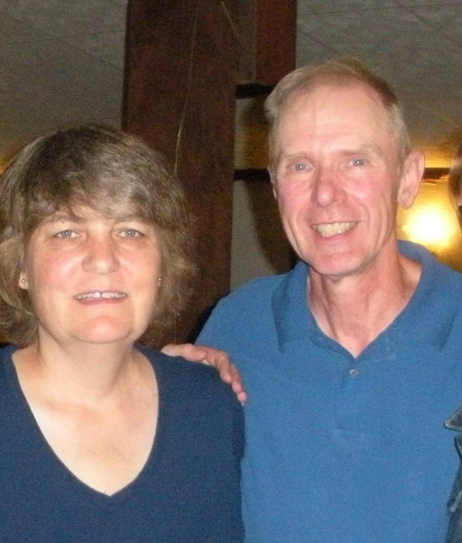 A middle-aged woman with short brown hair and a middle-aged man with short blond hair smiling, standing together indoors.
