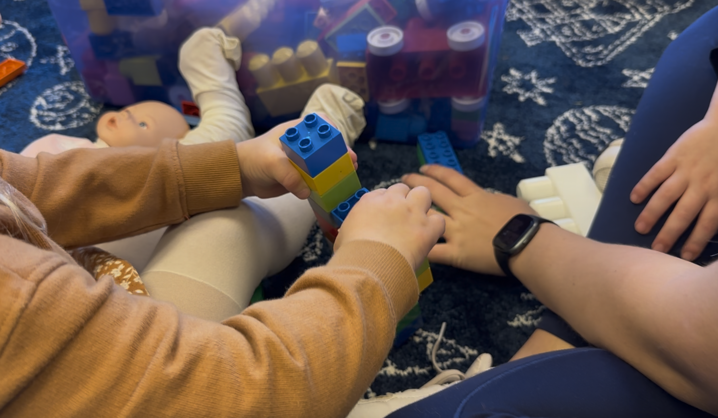 Children playing with colorful building blocks and a doll on a dark patterned carpet.