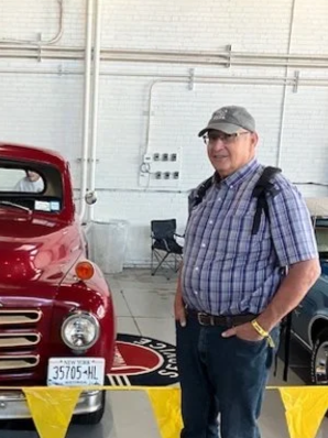 A smiling man in a plaid shirt, gray cap, and glasses stands inside an indoor space next to a vintage red car, with yellow caution tape around the car.