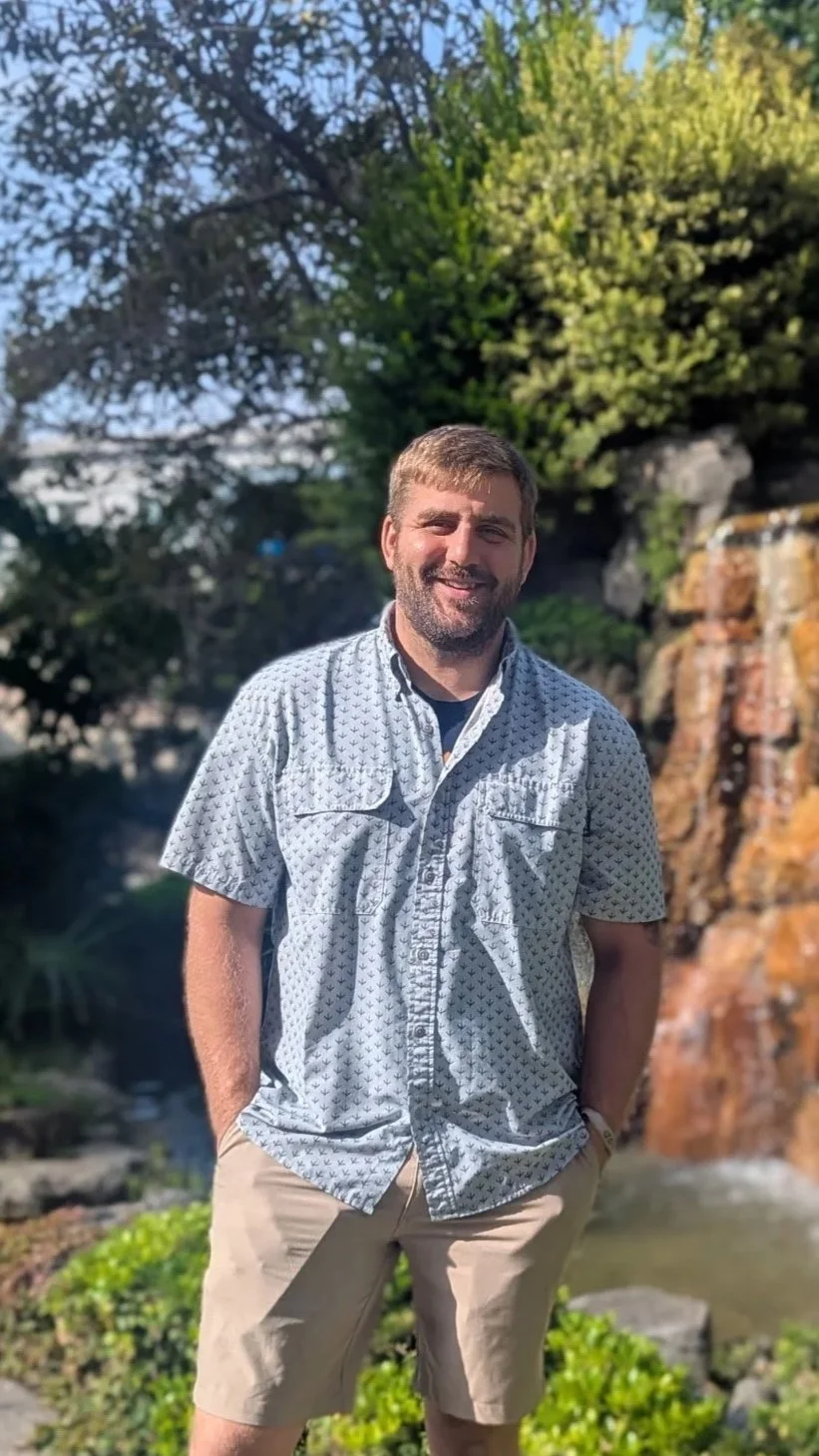 A man with short brown hair and a beard smiling outdoors, wearing a short-sleeved button-up shirt with a small pattern and light-colored shorts, standing in front of a waterfall and lush greenery.