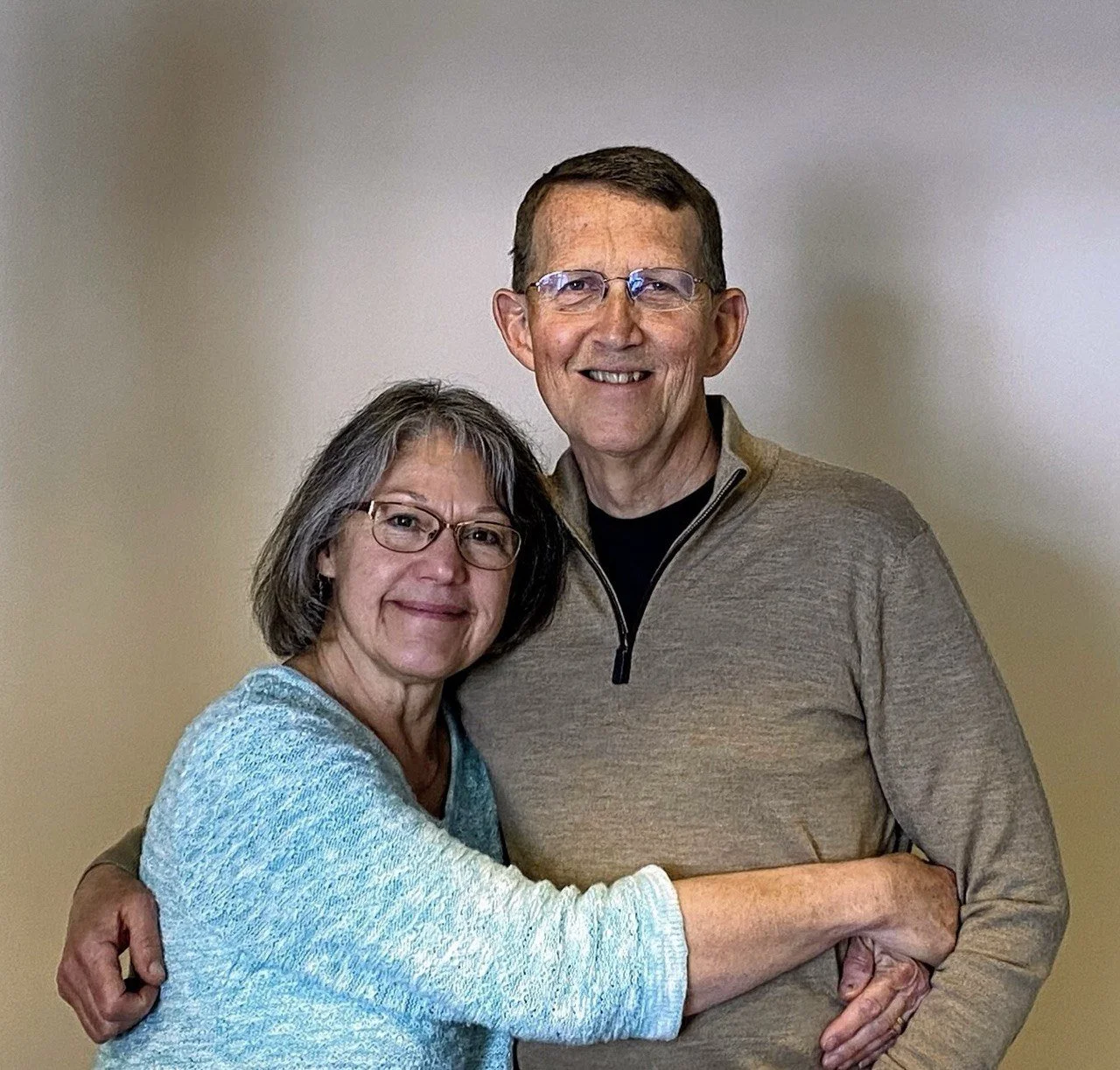 A smiling middle-aged couple hugging each other in front of a plain beige wall.