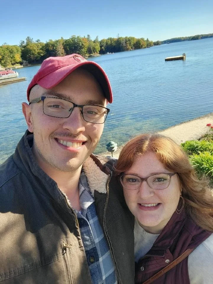 A smiling man wearing glasses, a red cap, and a jacket, along with a smiling woman wearing glasses and a maroon scarf, take a selfie by a lake with clear water, trees on the opposite shore, and a blue sky.