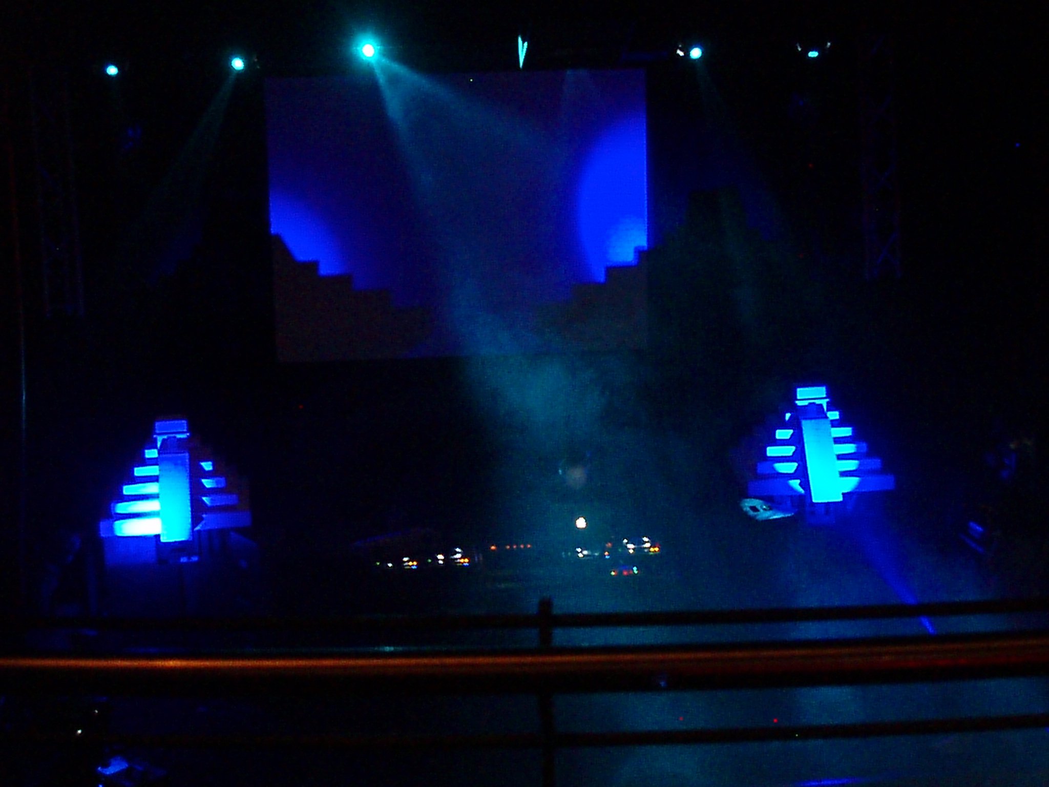 Dark stage with blue illuminated stairs and large central screen showing blue shapes, stage lights shining downward.
