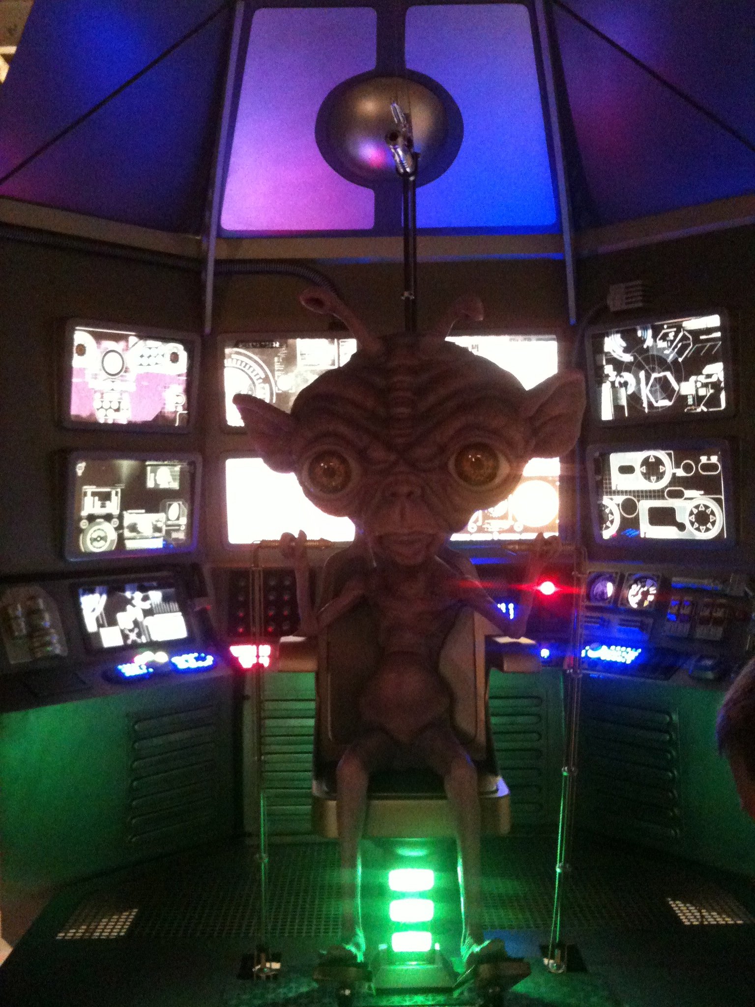 A model of E.T. the Extra-Terrestrial sitting in a spaceship command console with multiple monitors and control panels illuminated in a dark room.