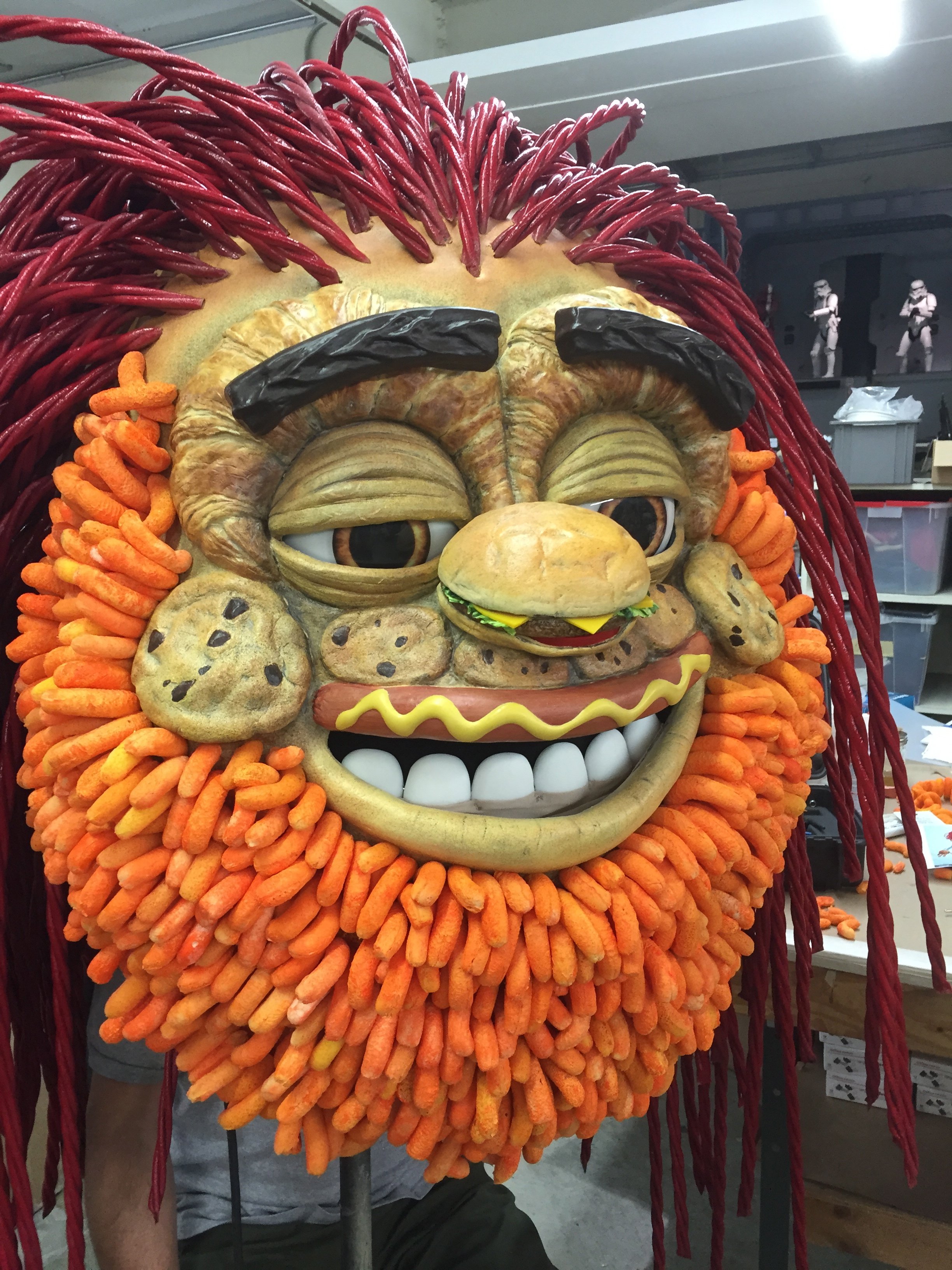 A large artistic mask resembling a cartoonish human face with exaggerated features, made from various snack foods like cookies, chips, and cheese puffs, with red licorice for hair and eyebrows.