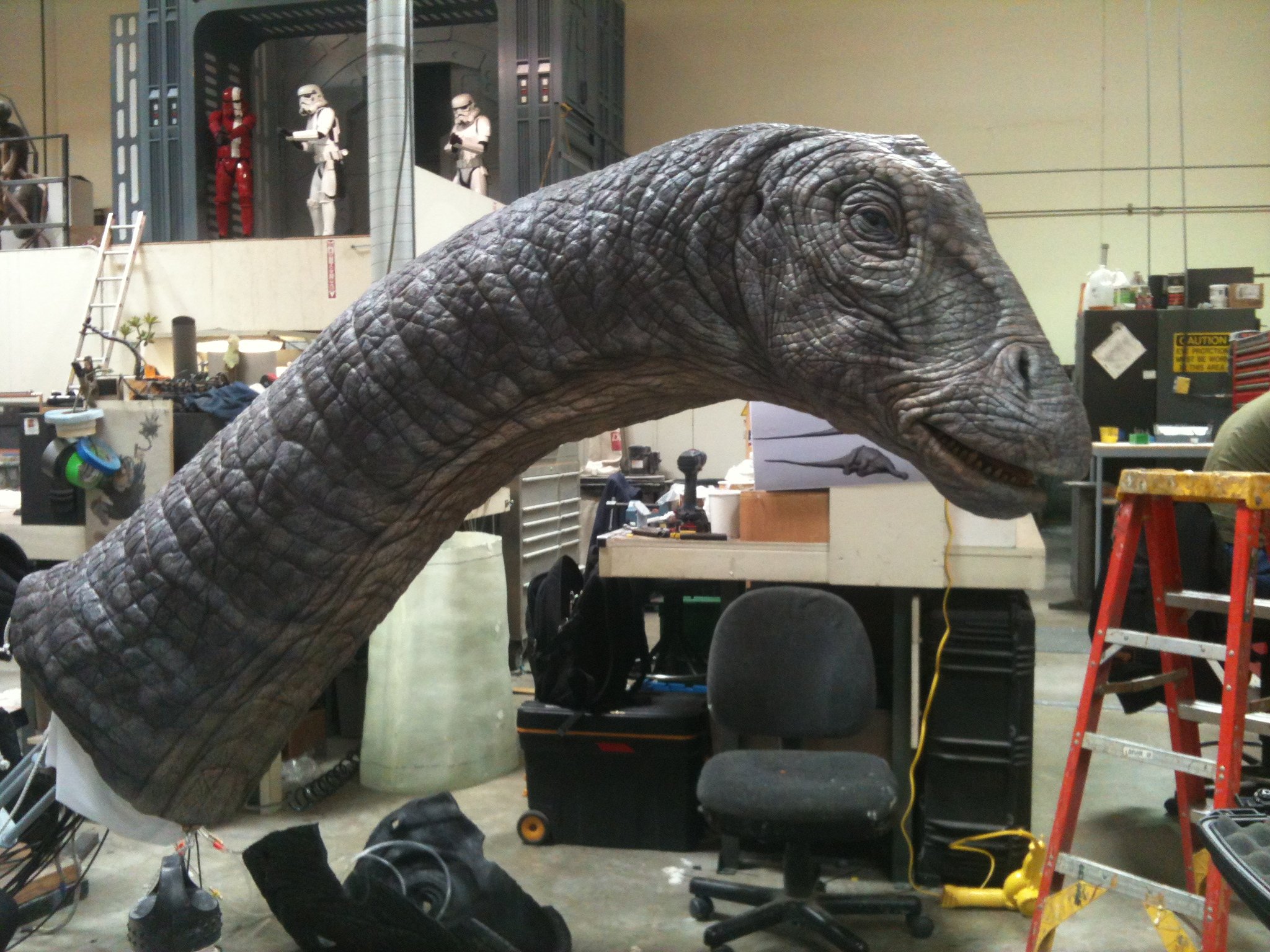 A person wearing a mask is behind a realistic dinosaur costume with a long neck, resembling a sauropod. The costume is in a workshop with tools, chairs, and a ladder.