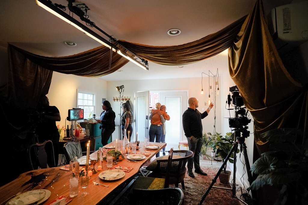 Behind-the-scenes view of a filming set in a dining room, with people preparing and adjusting lighting and camera equipment around a decorated dinner table.
