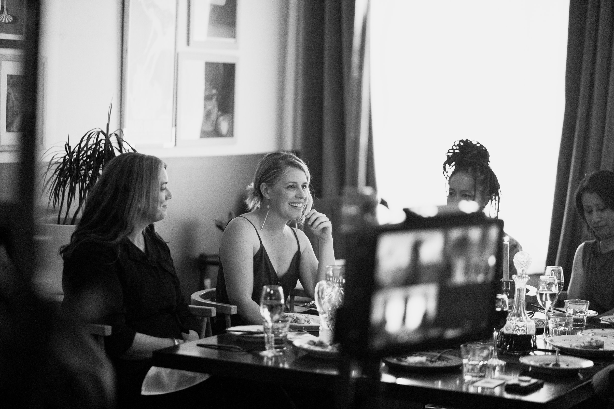 Black and white photo of women sitting at a dinner table, smiling and engaged in conversation, with plates, glasses, and a decanter on the table.