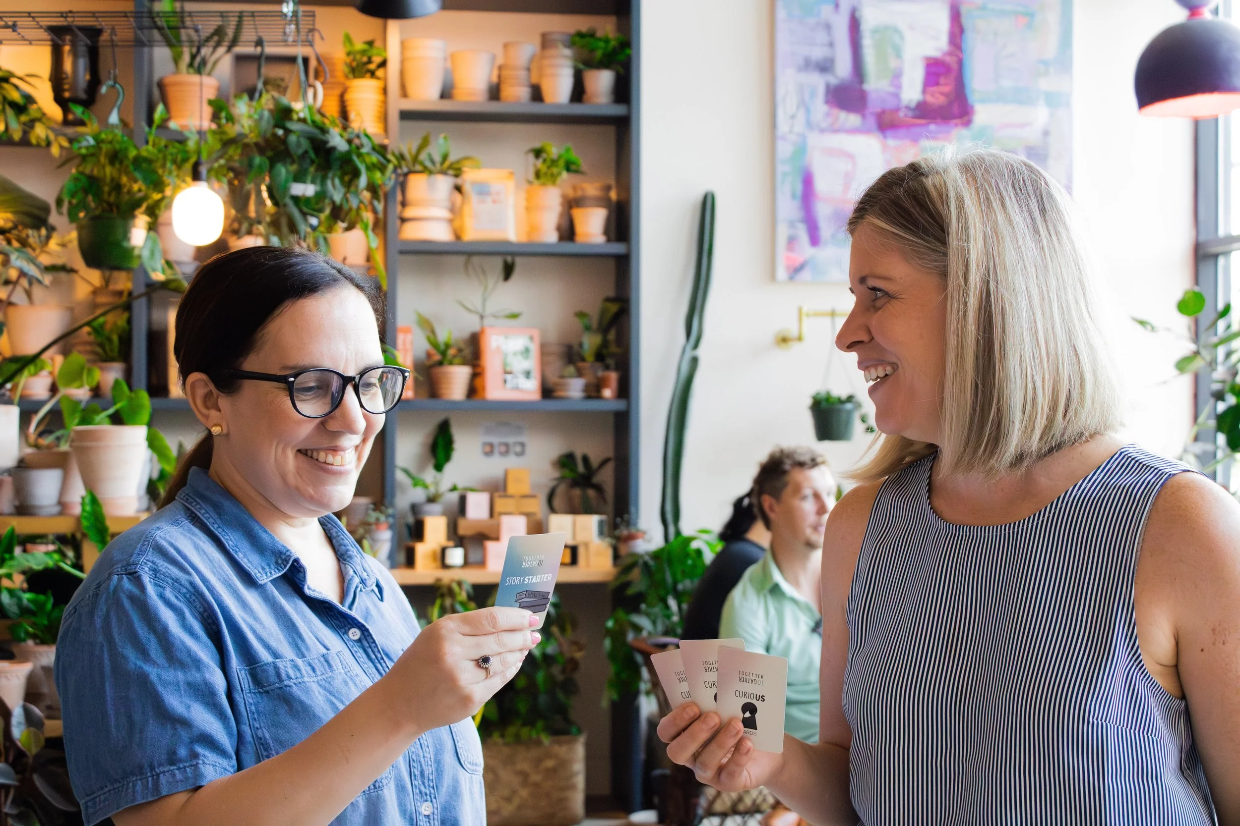 Two women smiling and playing a card game in a plant shop with shelves of potted plants and art on the walls.