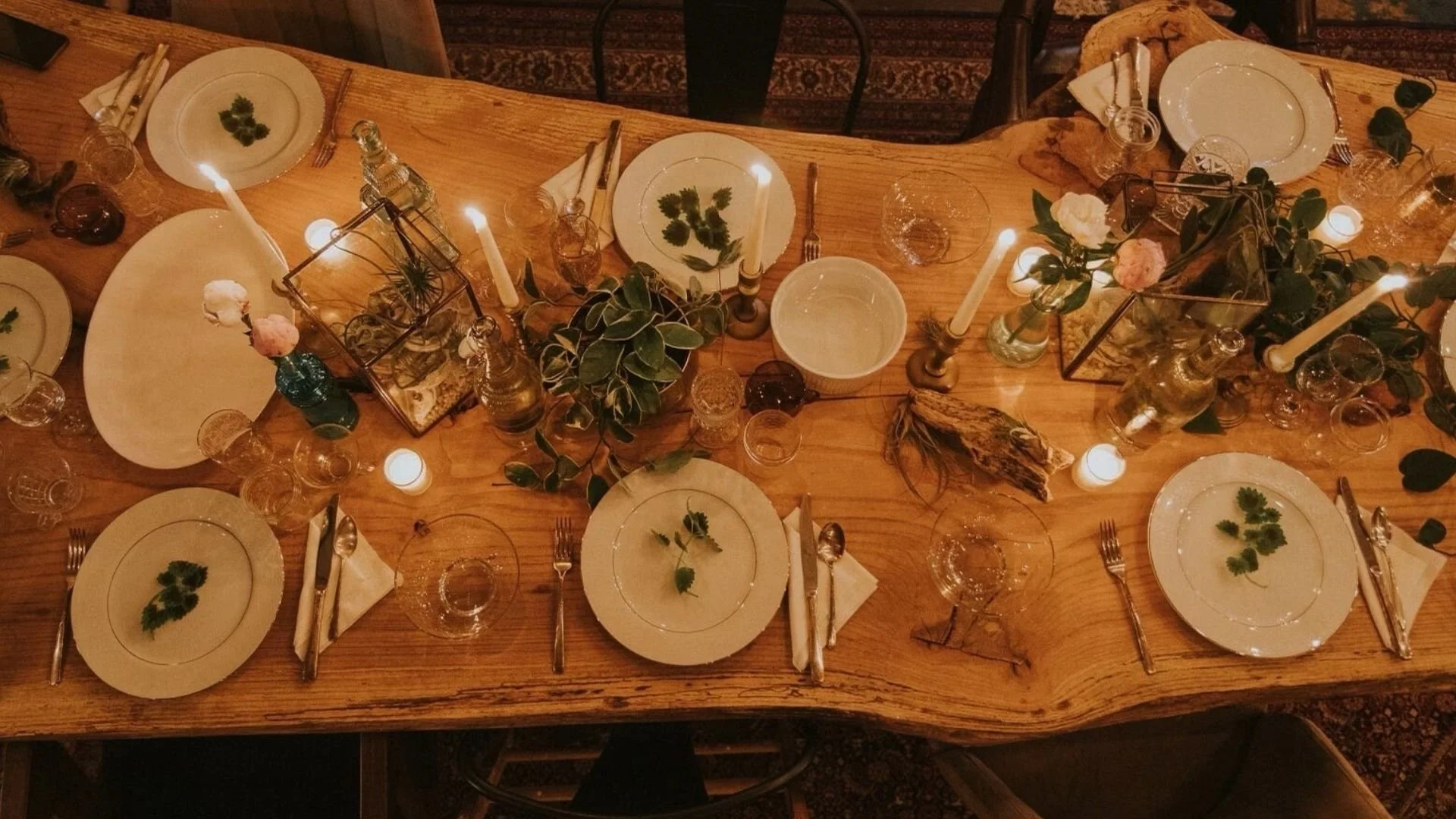 A top-down view of The Traveling Table set for a meal, with white plates decorated with green herbs, silverware wrapped in napkins, clear glasses, candles, greenery, and decorative elements inside glass containers.
