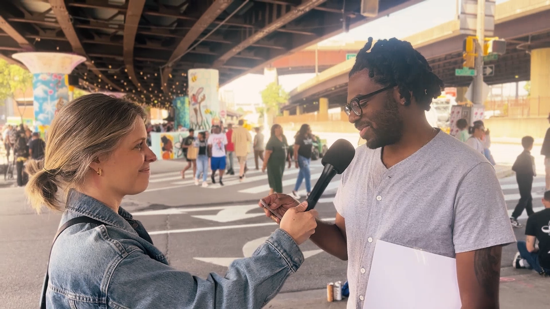 A woman wearing a denim jacket holds a microphone as she interviews a man on a city street under an overpass, with people walking in the background.