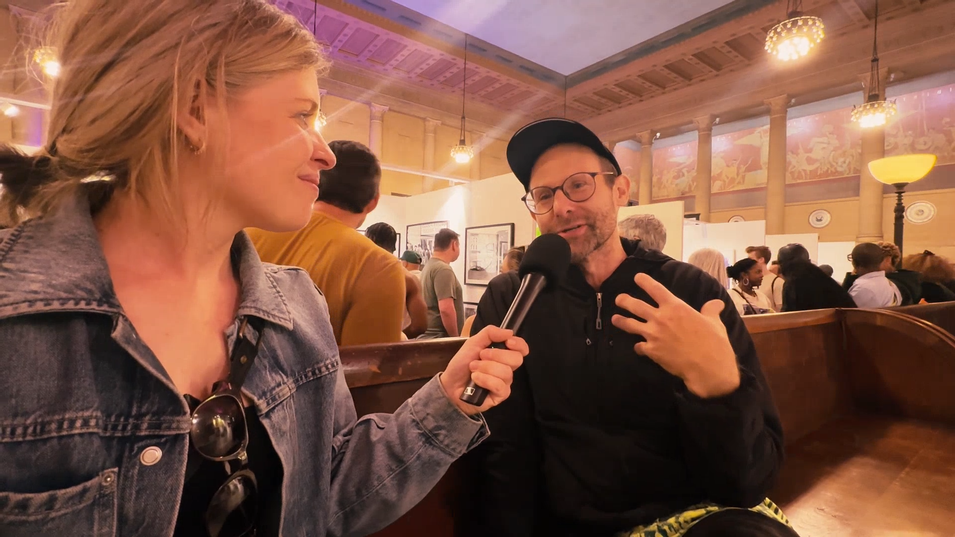 A woman with blonde hair in a ponytail holding a microphone, interviewing a man with glasses, beard, and wearing a black cap and black hoodie, seated in a crowded indoor space with warm lighting and artwork on the walls.