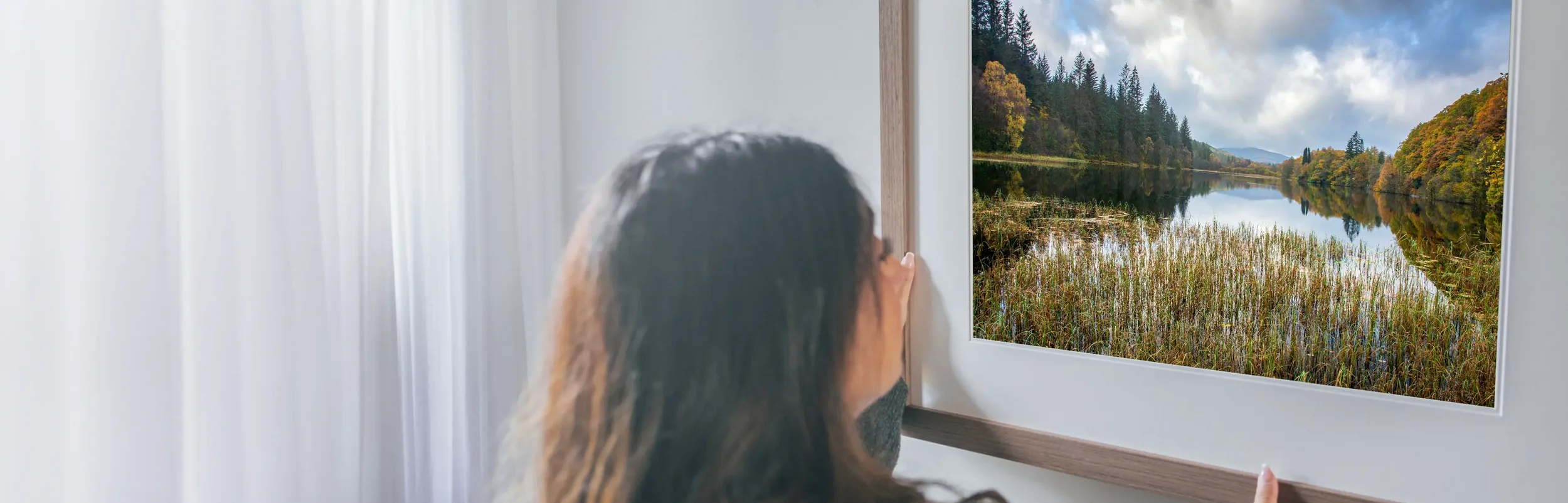 Person looking out of a window at a scenic lake and forested landscape with trees showing fall colors.