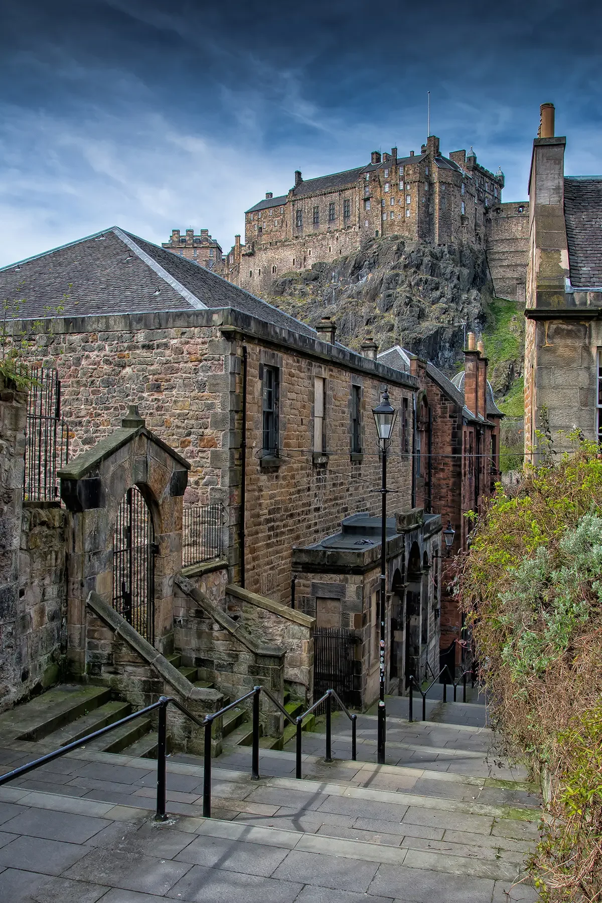 History-rich stone buildings with stairs and street lamps lead towards a large castle atop a rocky hill under a partly cloudy sky.