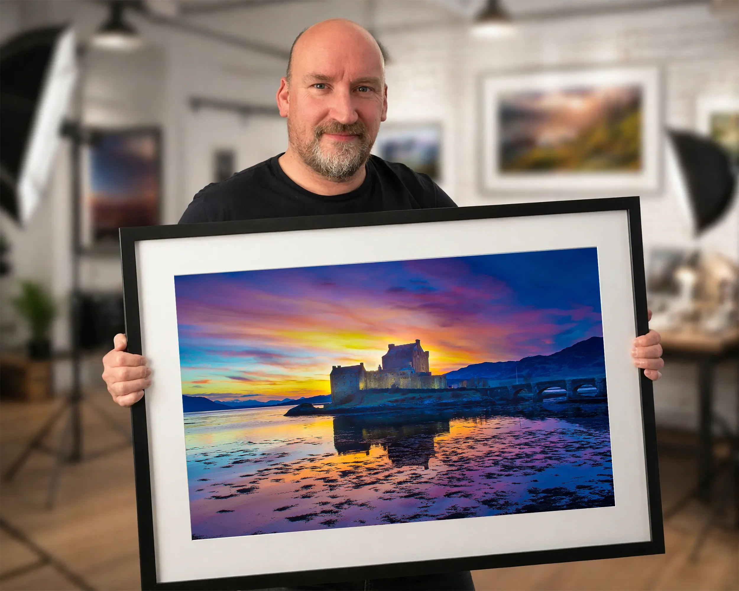 A man with a beard and mustache holding a framed colorful landscape photograph of a castle at sunset by a body of water with mountains in the background inside an art gallery or studio.