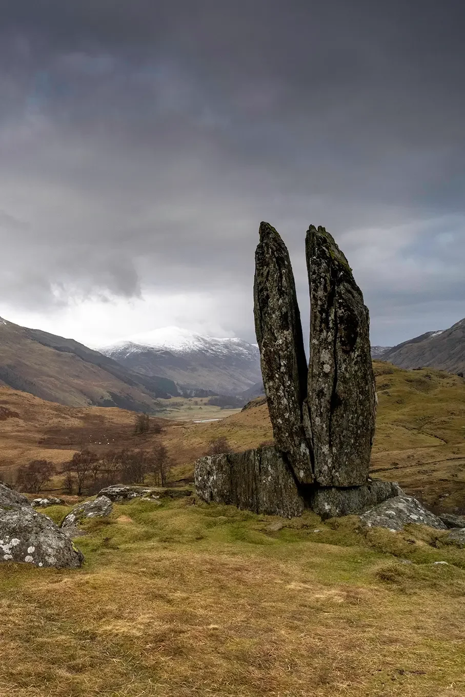 Large standing stone structure resembling fingers in a grassy landscape with hills, mountains with snow in the background, and dark overcast sky.