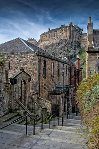Stone buildings with stairs and railing leading uphill towards a castle on a hill, under a blue sky with some clouds.
