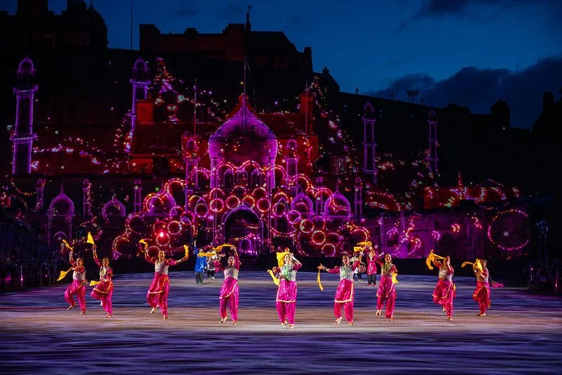 A group of performers dancing on stage in colorful costumes with a brightly lit castle backdrop during a nighttime show.