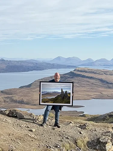Man standing on a rocky hill holding a large framed photograph that depicts the landscape behind him, with mountains and a body of water, under a partly cloudy sky.