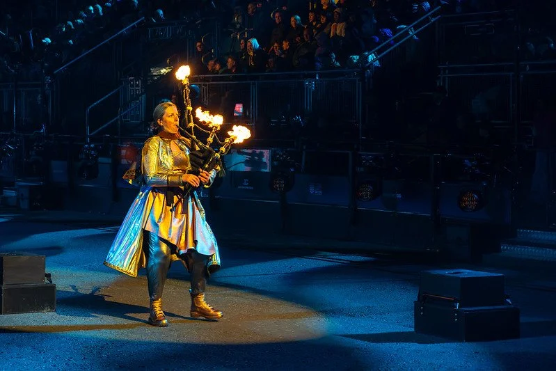 A performer dressed in a shiny costume holding a fire torch act in front of an audience in a dark, indoor arena.