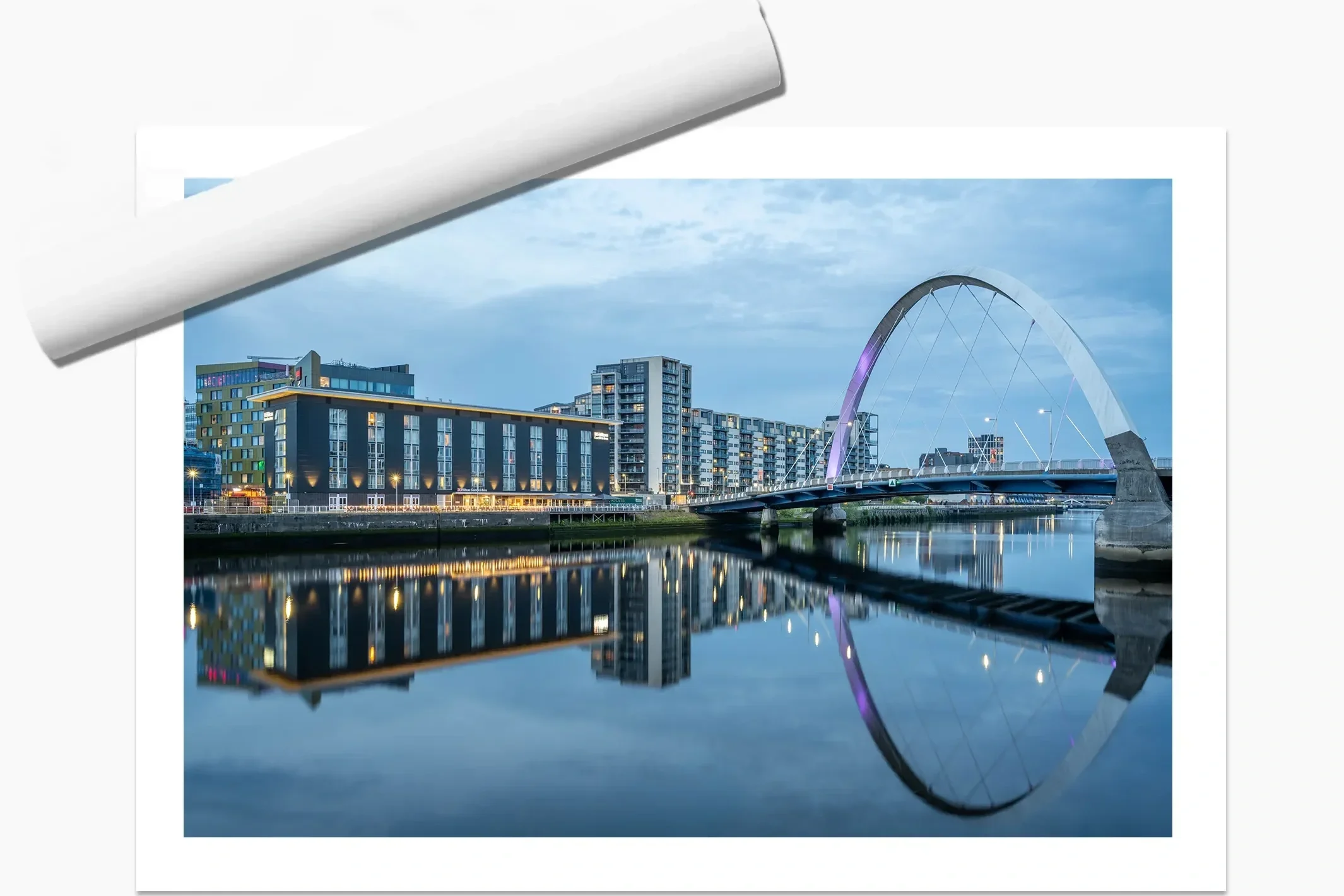 A cityscape at dusk with modern buildings and a bridge with an arch over a river, reflecting in the water below.