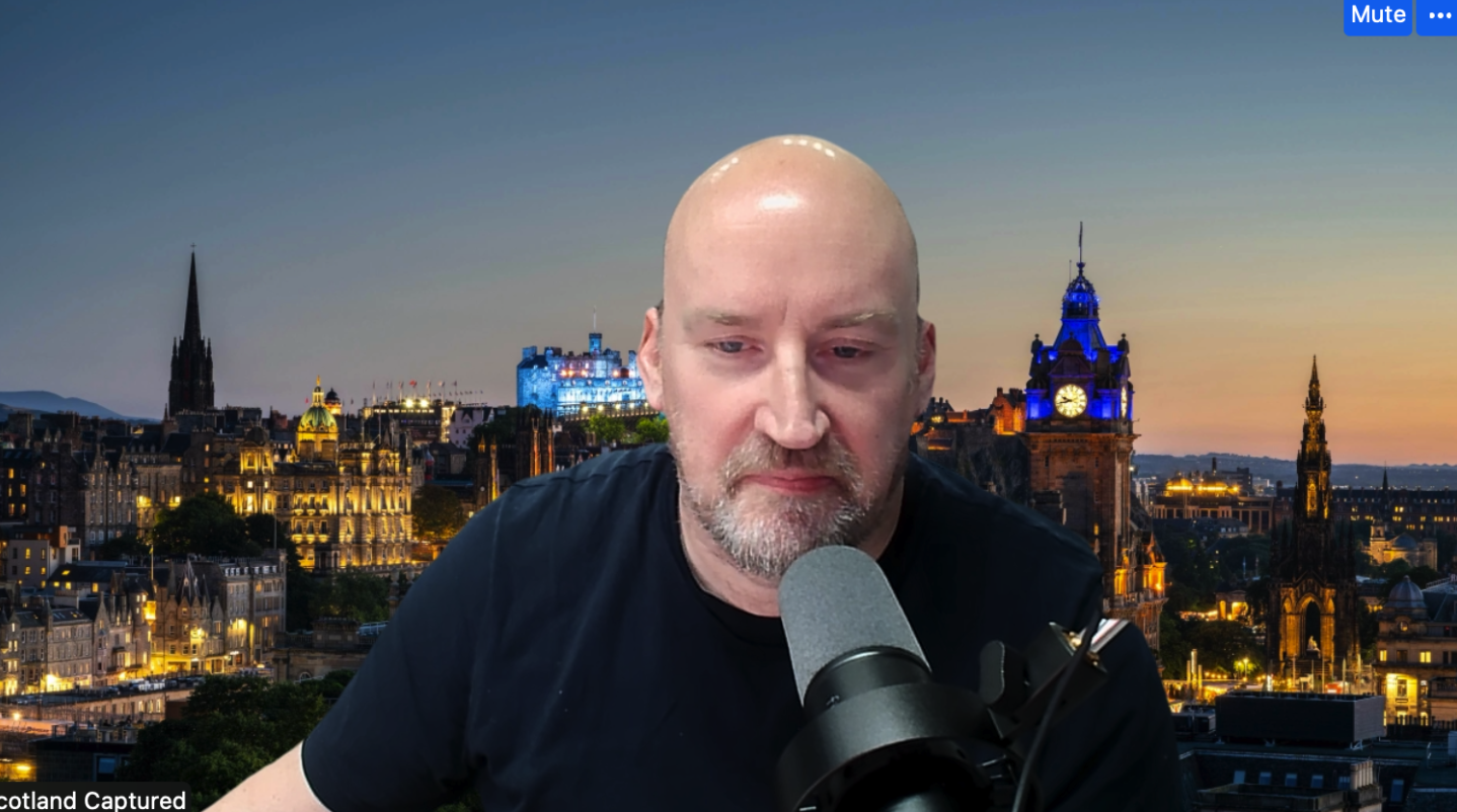 A man with a shaved head and gray beard sitting in front of a city skyline at sunset, speaking into a gray microphone.