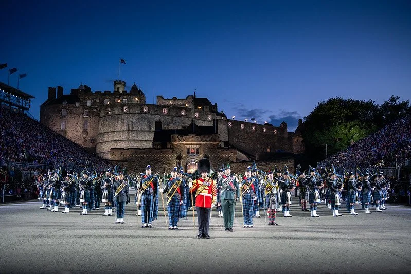 A large outdoor military parade or ceremony at night in front of a historic castle with marching soldiers in uniform, some with drums and swords, and spectators watching from the sides.
