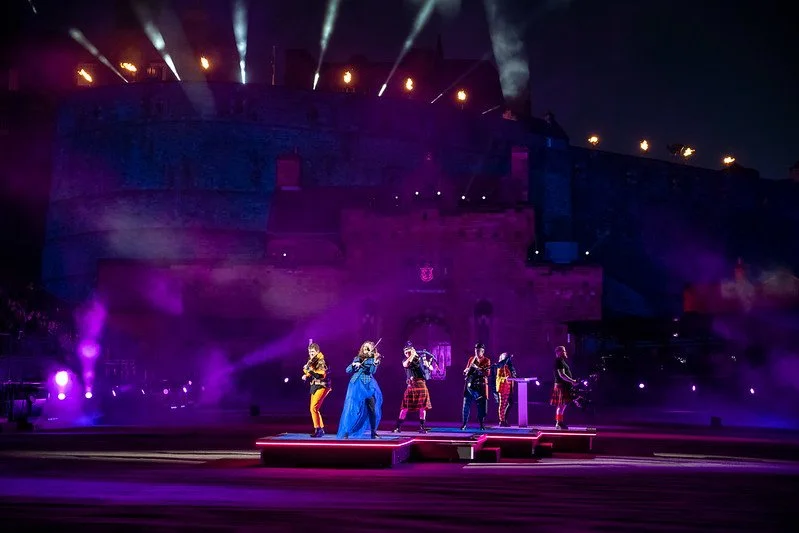 A live performance at a castle-themed venue with six performers on stage, illuminated by colorful lighting and smoke effects, at night.