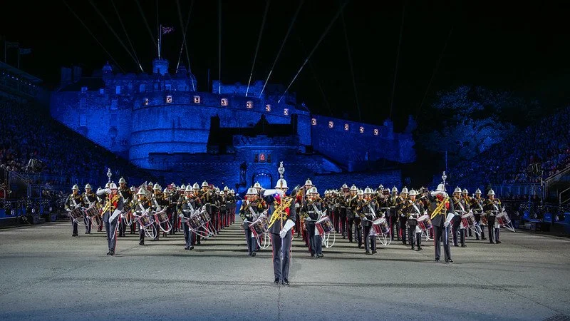 A marching band performing in front of a castle with blue lighting and laser beams at night, with an audience on either side.