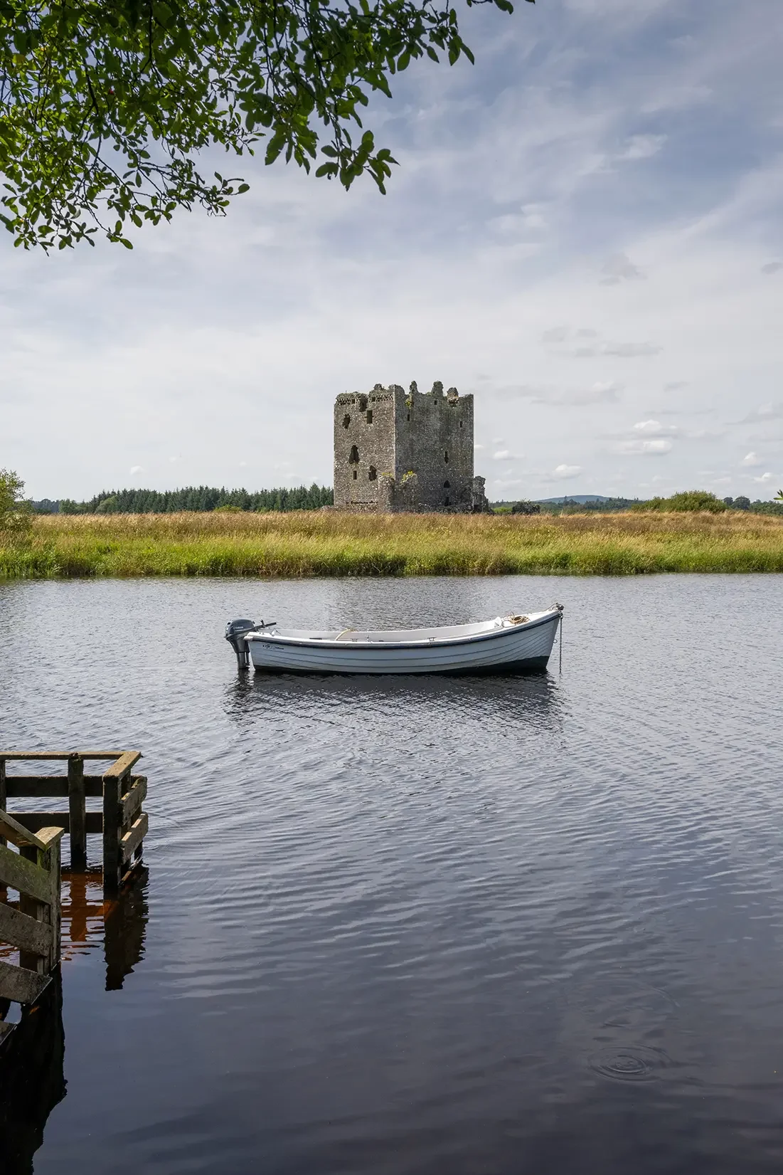 A small white boat floating on a calm body of water with a historic stone castle in the background and green trees along the horizon.