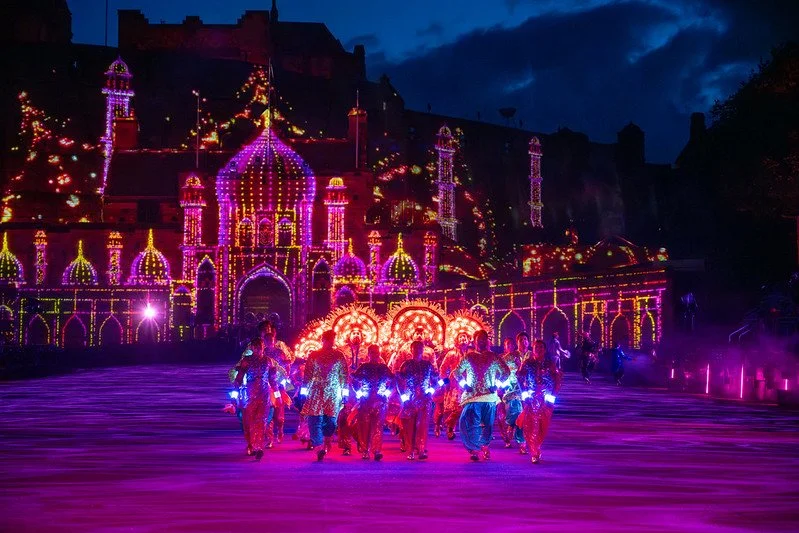 A group of people dressed in traditional attire walking towards a brightly lit, colorful building with domes and minarets, illuminated with neon lights at night.
