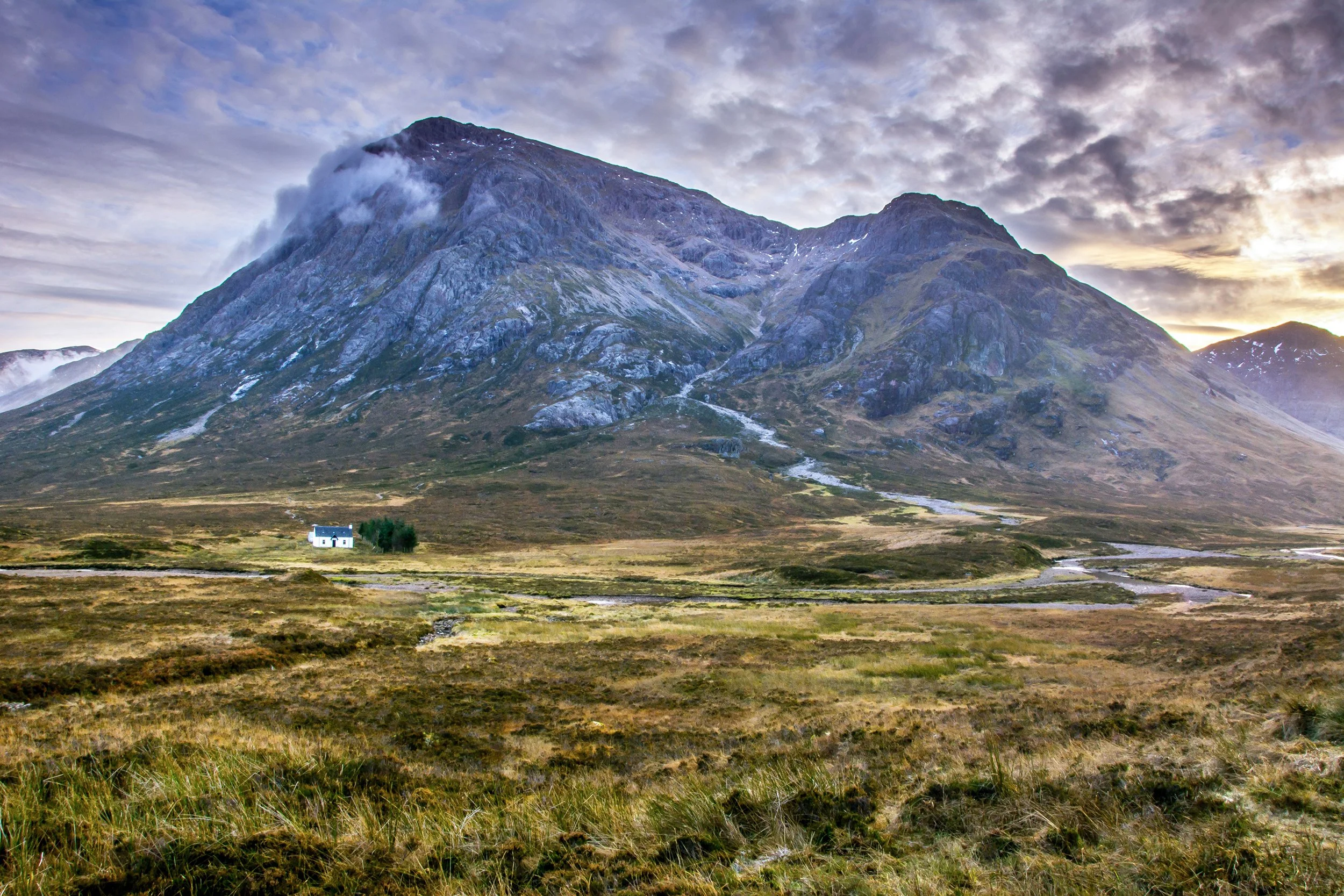 A solitary white house with a black roof in a green field, with a large mountain covered in patches of snow and clouds in the background during daytime.