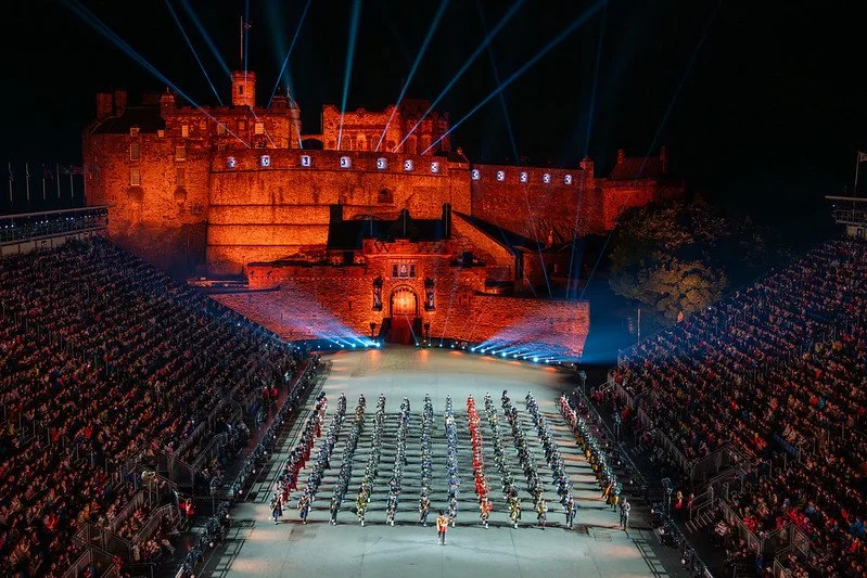 Large outdoor event at a historic castle illuminated by colorful lights, with rows of performers in uniform on a stage and a large audience watching.