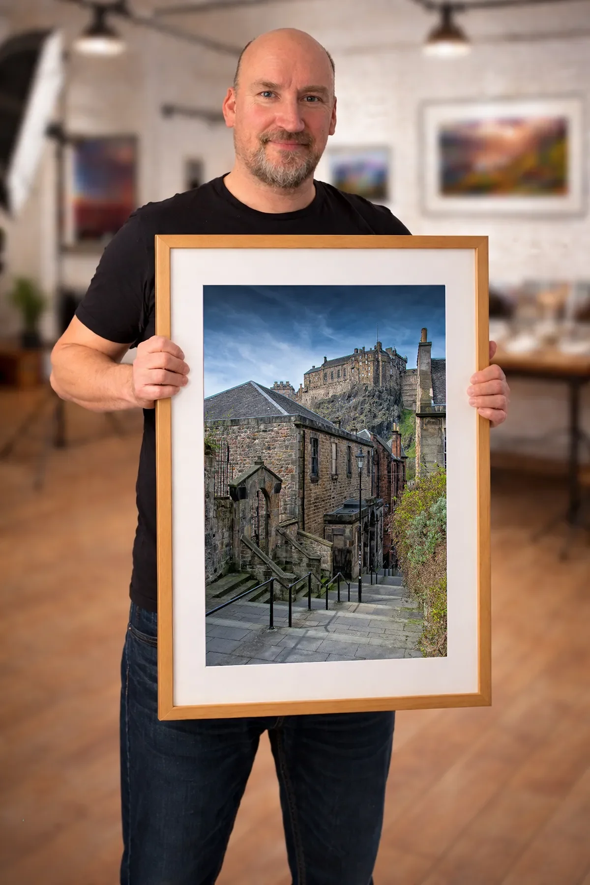 A man with a beard holding a framed photograph of a historic castle and stone buildings on a city street.