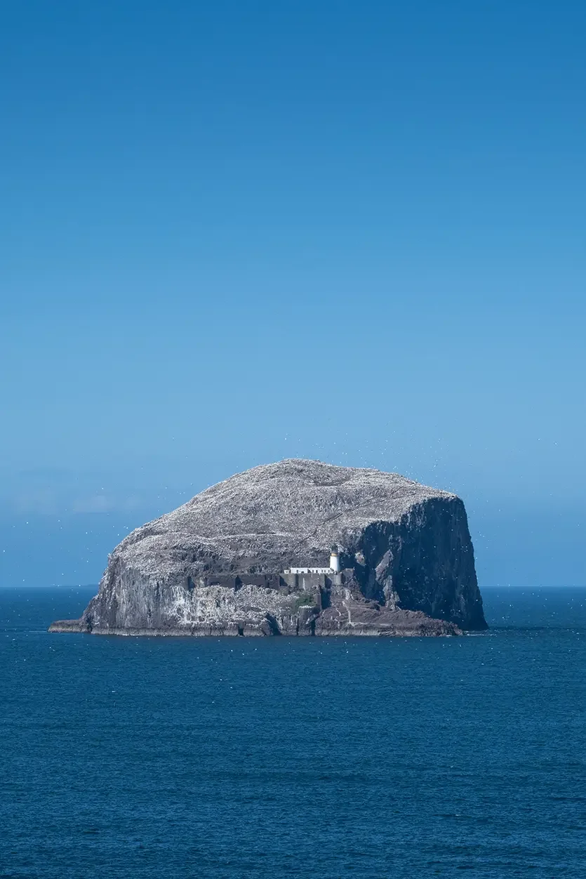 An island with a rocky, flat-topped summit surrounded by ocean water, with a lighthouse and some buildings near its base, under a clear blue sky.