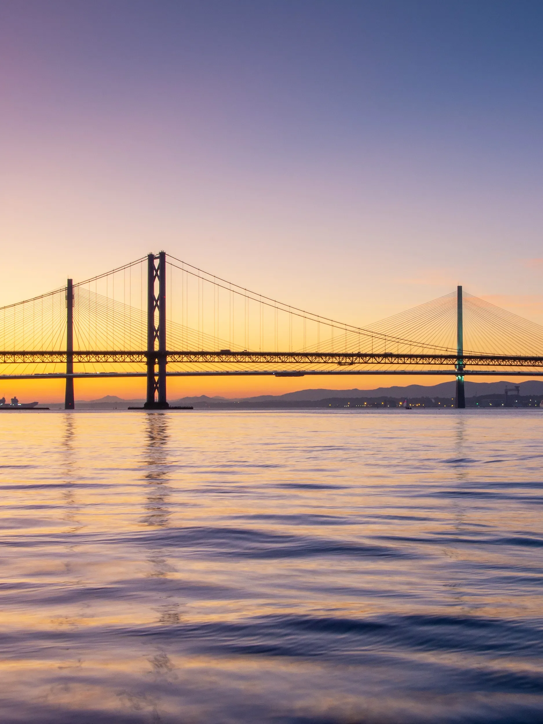 Silhouette of a large suspension bridge at sunset over a calm body of water.