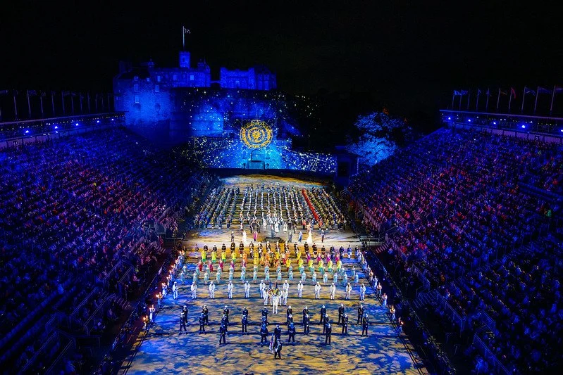 Nighttime scene at a large outdoor event with a castle in the background and a stage with performers, surrounded by large crowds on both sides.