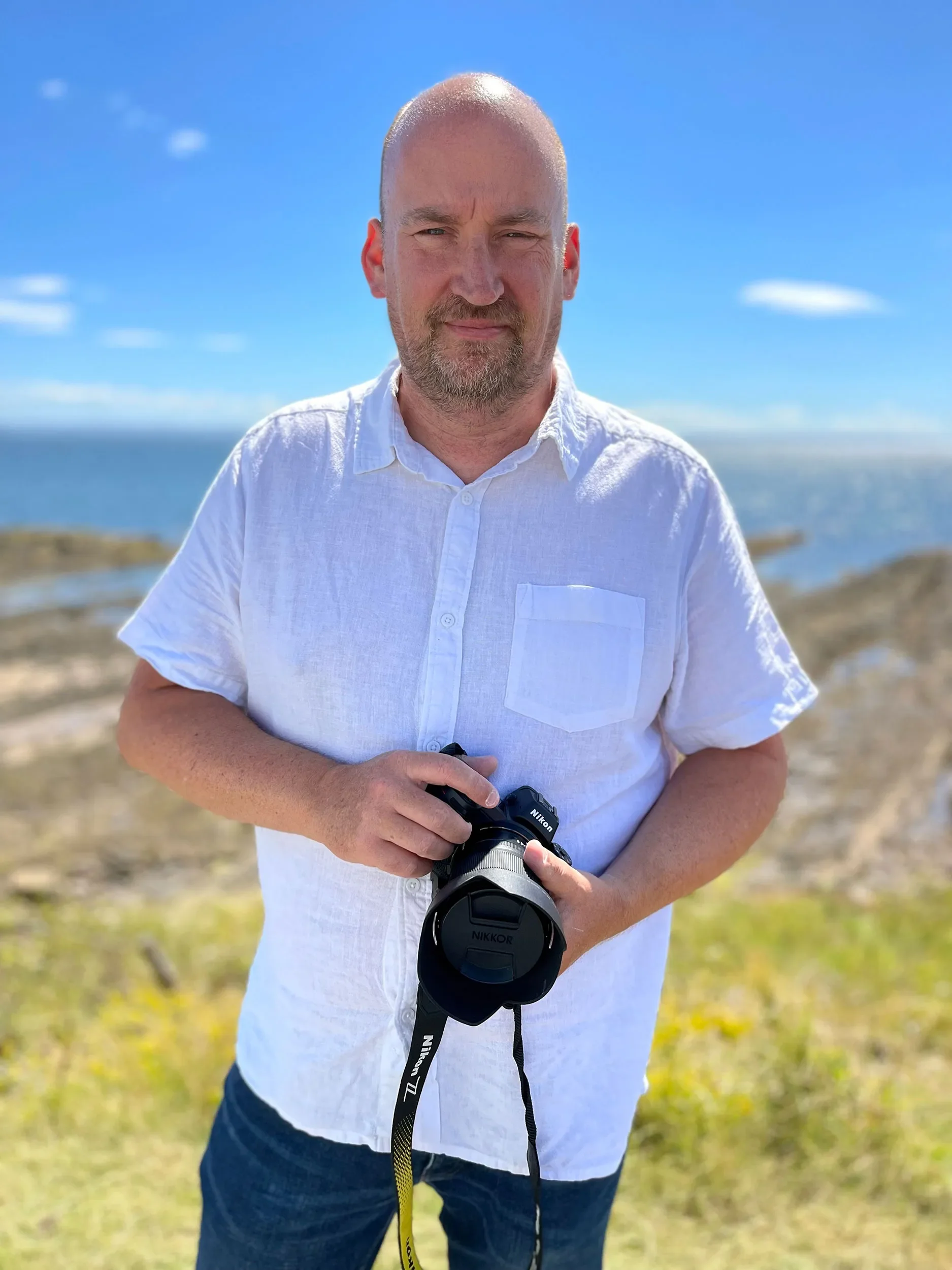 A man with a bald head, beard, and white shirt standing outdoors on a sunny day, holding a Nikon camera.