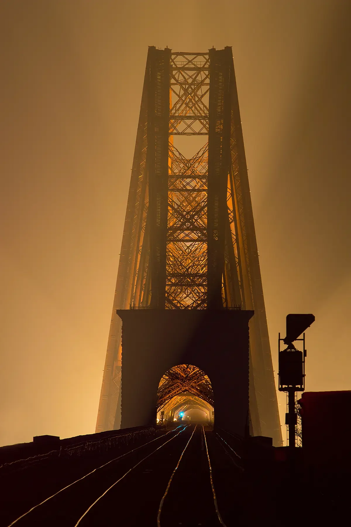 The image shows the Golden Gate Bridge's tower shrouded in fog at night, with train tracks leading towards it and a silhouette signal on the right side.